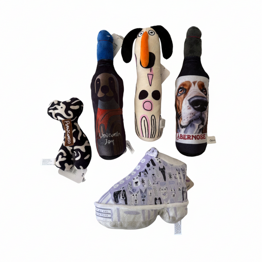 Five dog toys arranged on a plain white background: a black-and-white patterned bone-shaped plush, a dark bottle-shaped plush with a dog face and red label, a cream bird-shaped plush with black ears and an orange beak, a dark beer-bottle-shaped plush with a dog face and 'ABERNOSE' text, and a light purple shoe-shaped plush with dog illustrations. Small sewn tags are visible on some items.